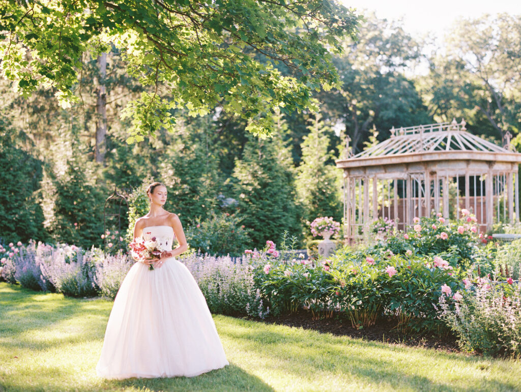 A bride in a strapless Lein Studio gown stands gracefully in a blooming garden at Greencrest Manor, with a vintage glass conservatory in the background, captured on film.