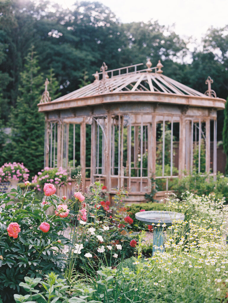 A vintage conservatory surrounded by lush garden blooms, including vibrant pink peonies and delicate wildflowers, at Greencrest Manor, captured on film.