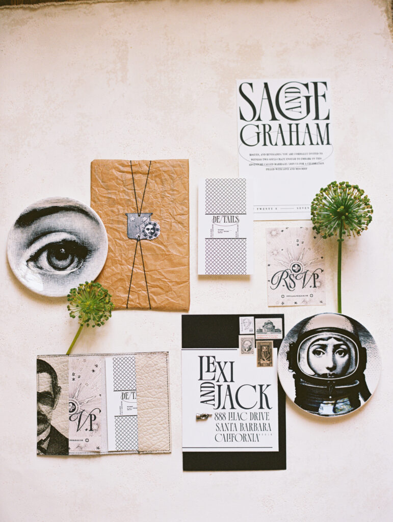 A film photography flatlay of modern wedding stationery featuring bold typography, artistic illustrations, vintage stamps, and textured paper details, styled with sculptural floral elements.