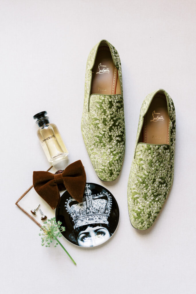 Groom’s wedding details featuring green embroidered Christian Louboutin loafers, a brown velvet bow tie, luxury cologne, and elegant cufflinks, captured by Lexie Vaccaro Photography at Greencrest Manor.