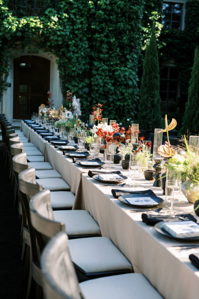 A long, elegantly set outdoor dining table at Greencrest Manor, featuring black tableware, sculptural floral arrangements, and warm candlelight, all framed by lush ivy-covered walls.