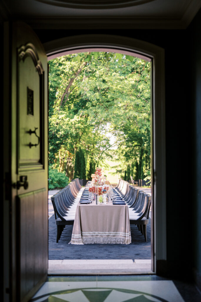 A beautifully set outdoor reception table at Greencrest Manor, framed by an open doorway. The long table is adorned with delicate florals and elegant tableware, nestled beneath lush green trees.