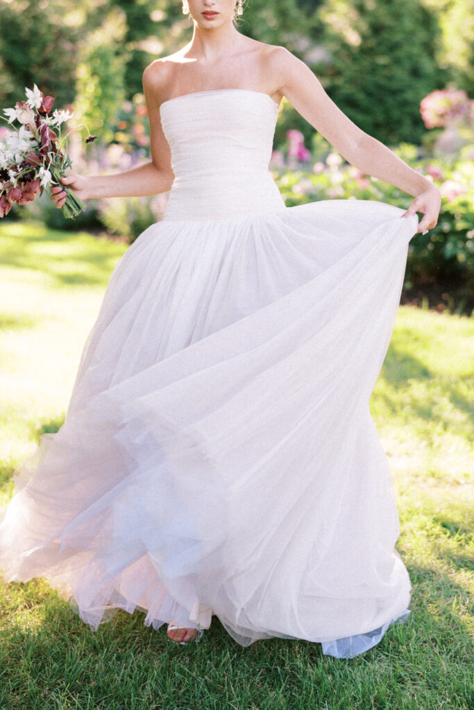 A bride gracefully lifts the flowing layers of her gown while holding a bouquet, bathed in golden sunlight at Greencrest Manor.