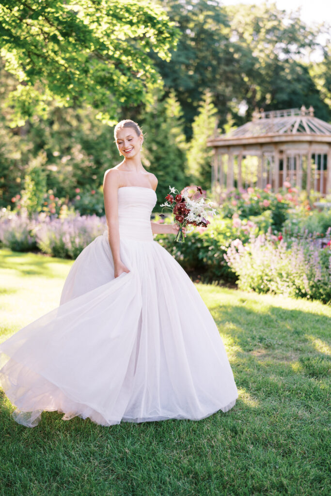 A radiant bride twirls in the garden at Greencrest Manor, her ethereal gown catching the sunlight as she smiles, holding a bouquet of deep red and white flowers.