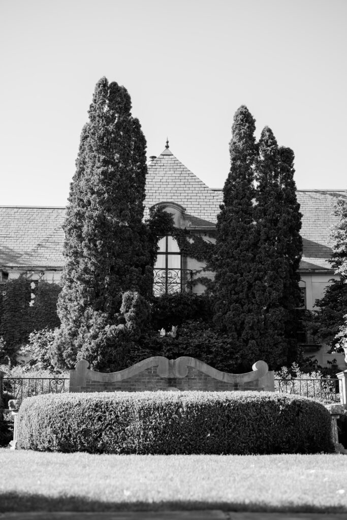 A black and white film photograph of Greencrest Manor, showcasing its European-inspired architecture, ivy-covered walls, and towering cypress trees. A sculptural stone bench sits in the foreground, adding to the estate’s old-world charm.