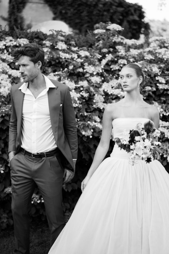 A black-and-white editorial portrait of a bride in a strapless Lein Studio gown and a groom in a tailored suit, walking past a lush floral backdrop at Greencrest Manor.