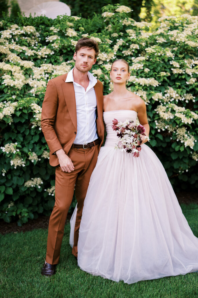 A fashion-forward bride and groom pose against a lush floral backdrop at Greencrest Manor. The bride wears a strapless Lein Studio gown and carries a sculptural bouquet, while the groom dons a rust-colored suit, exuding modern European elegance.