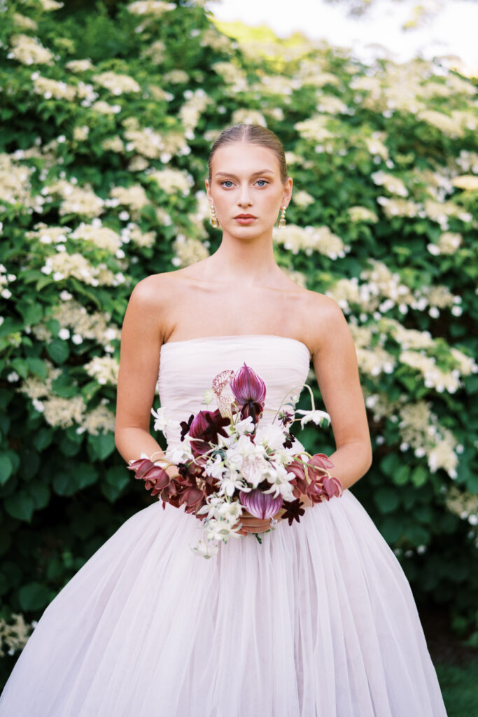 A bride in a strapless Lein Studio gown holds a sculptural bouquet in deep burgundy and ivory tones, standing against a backdrop of blooming white hydrangeas at Greencrest Manor.