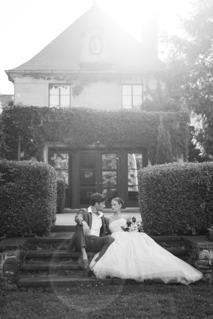 Black and white film photograph of a bride and groom sitting on stone steps outside a grand ivy-covered estate, bathed in soft sunlight.