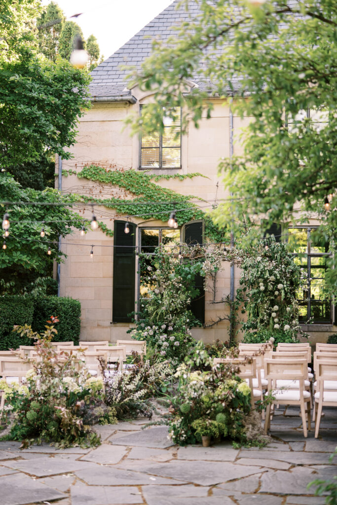 Enchanting outdoor wedding ceremony setup at Greencrest Manor with lush floral arrangements, elegant wooden chairs, and string lights, captured by Lexie Vaccaro Photography.