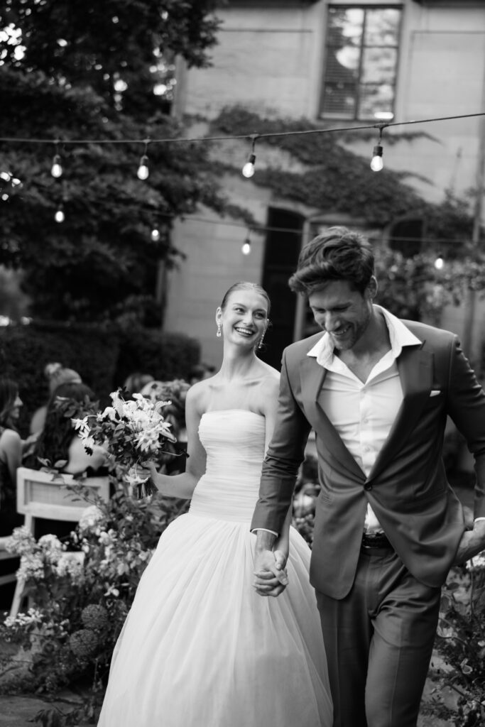 Black and white film photograph of a joyful bride and groom walking hand-in-hand down the aisle, surrounded by guests and glowing string lights.