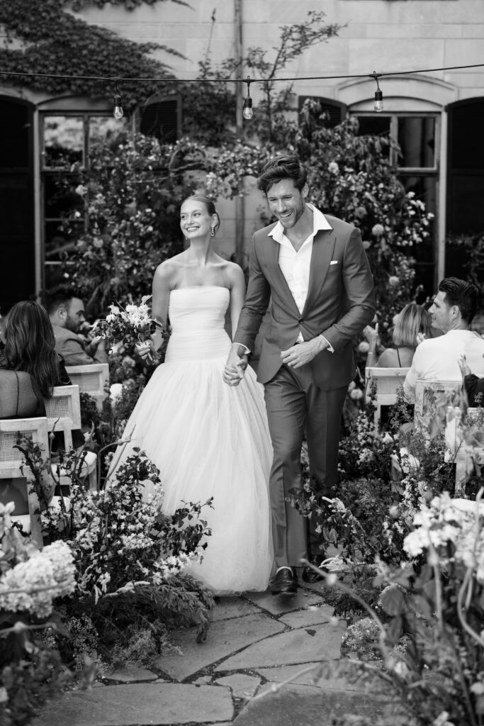 A black-and-white image of a bride and groom walking hand in hand down the aisle at Greencrest Manor, surrounded by lush floral arrangements and an intimate gathering of guests. The bride beams with joy as she carries her bouquet, and the groom smiles warmly in a tailored suit.