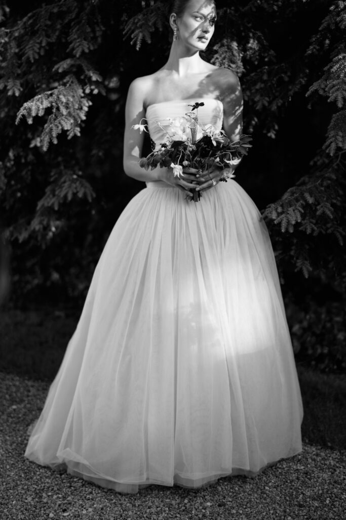 A black-and-white film portrait of a bride in a strapless Lein Studio gown, standing in dappled sunlight, holding a sculptural bouquet against a backdrop of lush greenery at Greencrest Manor.