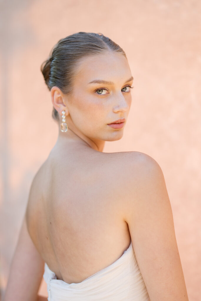 A soft portrait of a bride in a strapless gown, gazing over her shoulder with effortless elegance. Warm sunlight highlights her dewy skin and delicate features, with a neutral-toned Chasing Stone backdrop enhancing the ethereal glow.