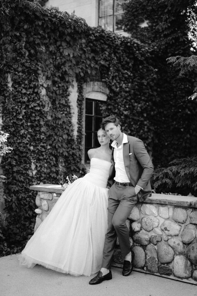 A black-and-white portrait of a bride and groom leaning against a stone wall, surrounded by lush ivy at Greencrest Manor. The bride wears a voluminous gown, and the groom dons a tailored brown suit with an open-collared white shirt.