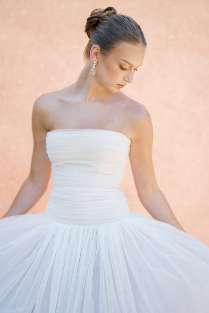 A bride in a strapless Lein Studio gown, softly looking down as golden light illuminates her delicate features. The voluminous tulle skirt billows gently, framed against a Chasing Stone backdrop.