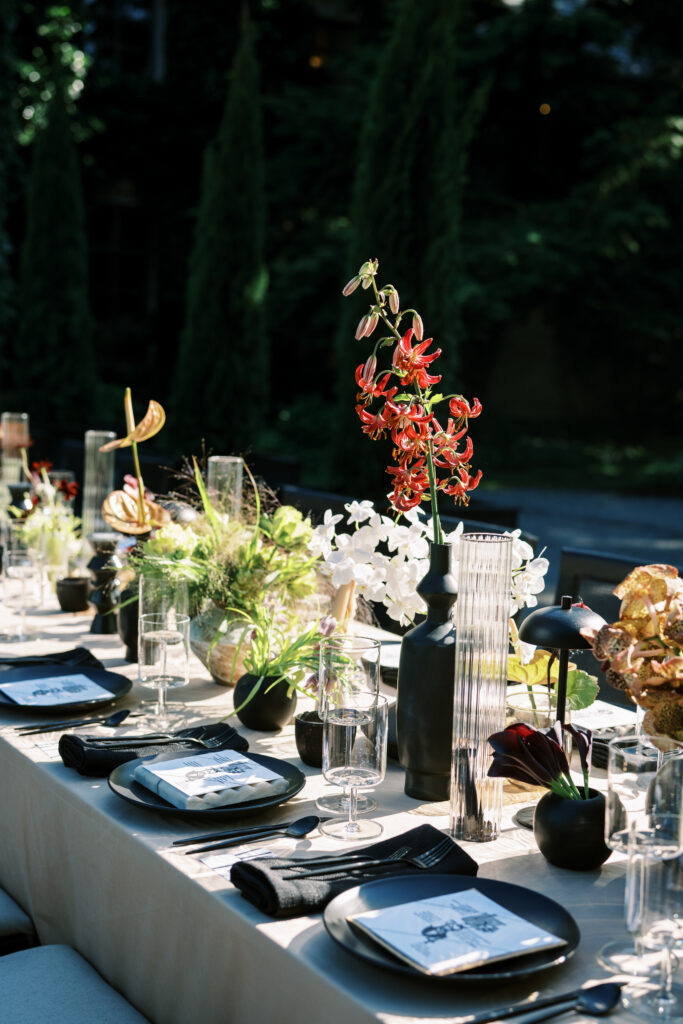 A dramatic outdoor tablescape at Greencrest Manor, featuring modern black tableware, sculptural floral arrangements, and a striking red lily centerpiece in the golden evening light.