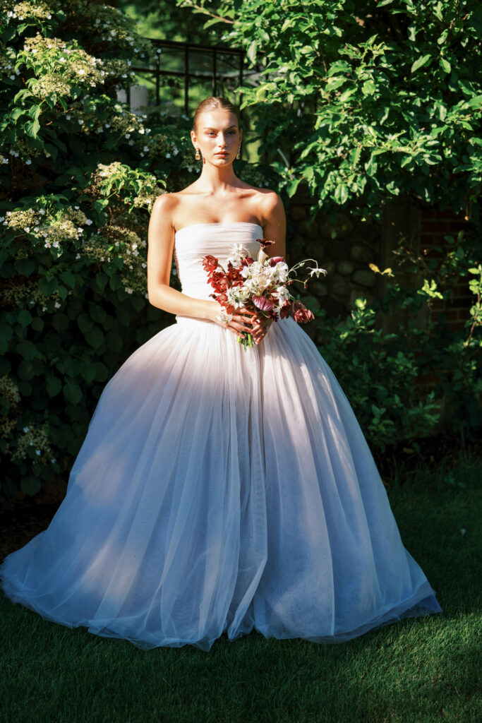 A bride in a strapless Lein Studio gown stands in dramatic golden light, holding a sculptural bouquet in deep red and ivory tones, against lush garden greenery at Greencrest Manor.