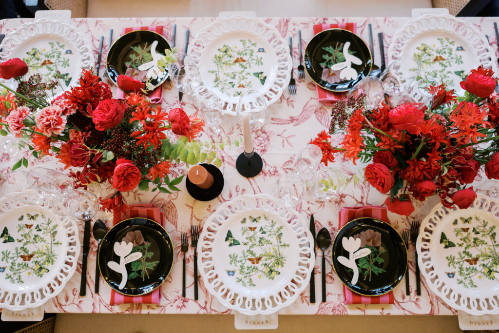 Overhead view of a wedding reception table with red and pink florals, butterfly-illustrated plates, black flatware, and pink napkins