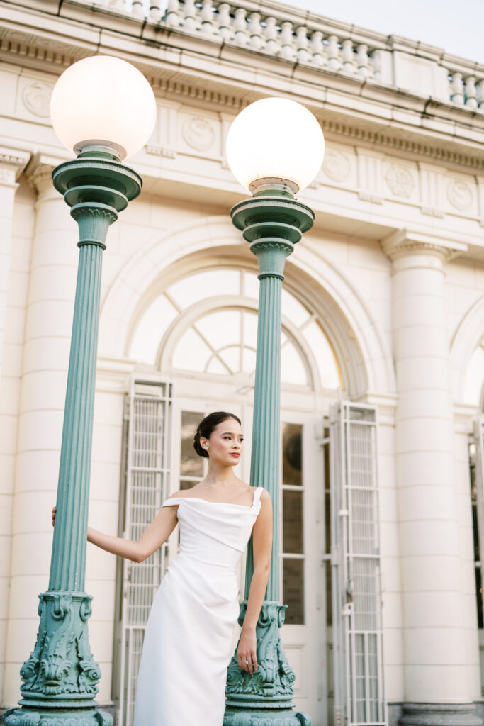 Bride in a modern white gown poses confidently between vintage teal lampposts at the Prospect Park Boathouse, photographed by Lexie Vaccaro Photography.

