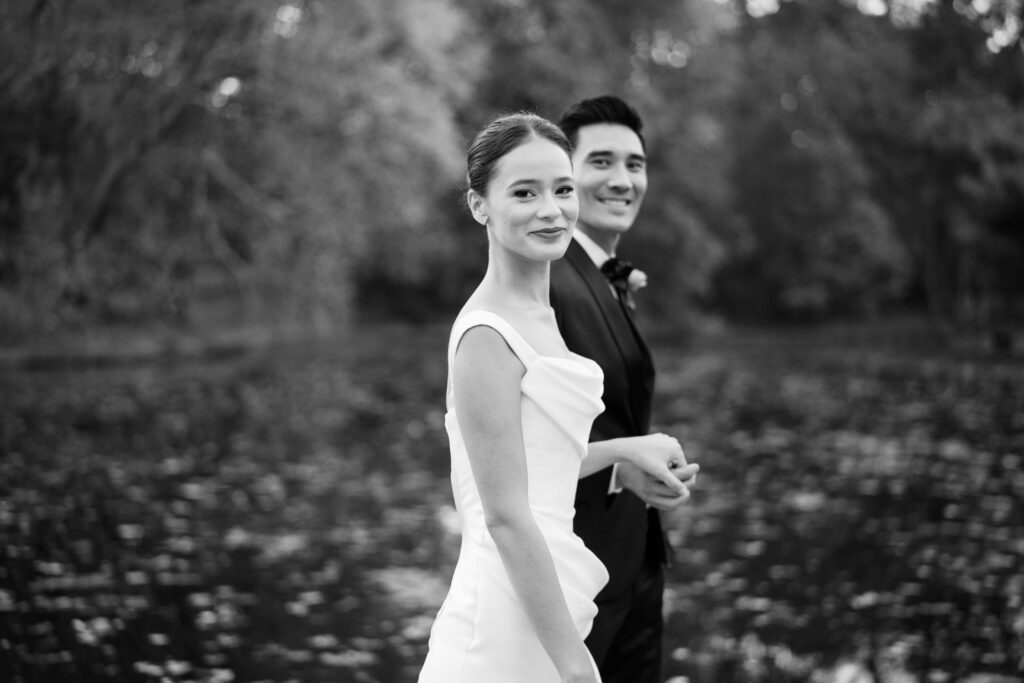 Black and white photo of bride and groom walking by the water, smiling at the camera
