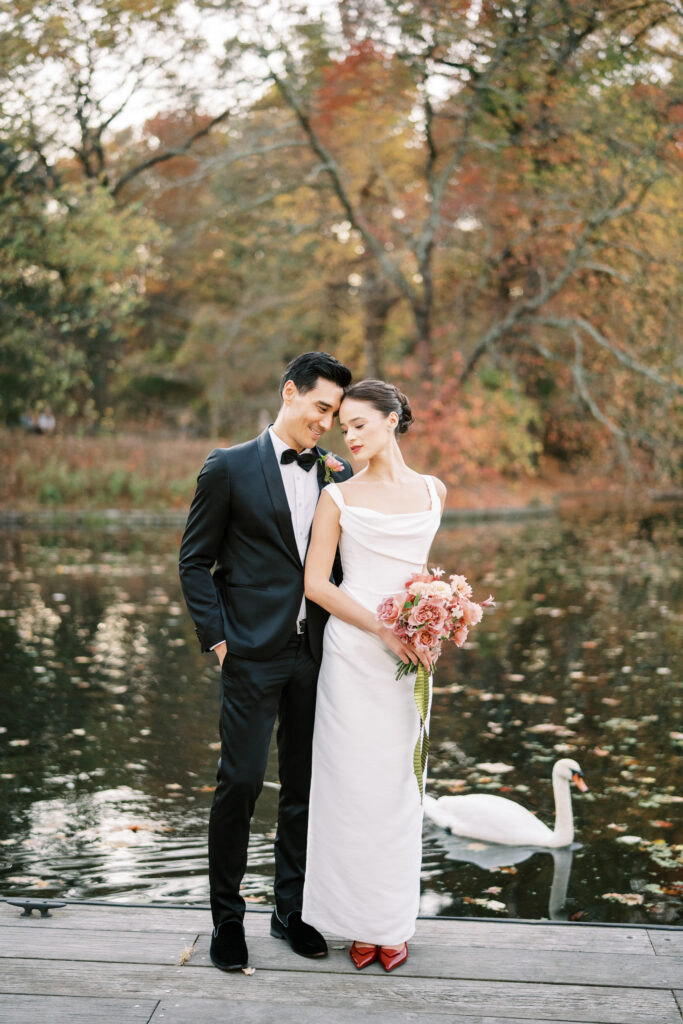 Bride and groom standing close together on a dock in front of a pond with a swan swimming nearby, surrounded by autumn trees