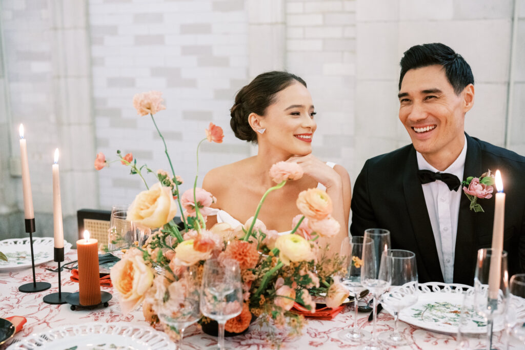 Bride and groom laughing at their candlelit wedding dinner surrounded by floral centerpieces.