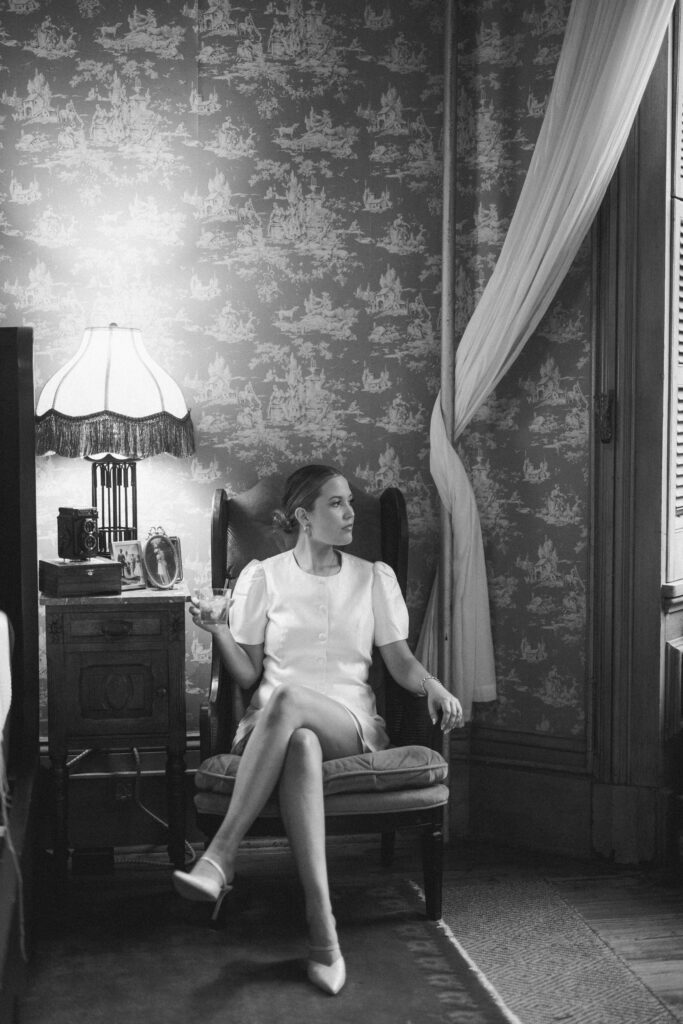 Black-and-white photo of Lexie Vaccaro seated in a vintage armchair with crossed legs, holding a drink and gazing toward a window beside a fringed lamp and antique side table