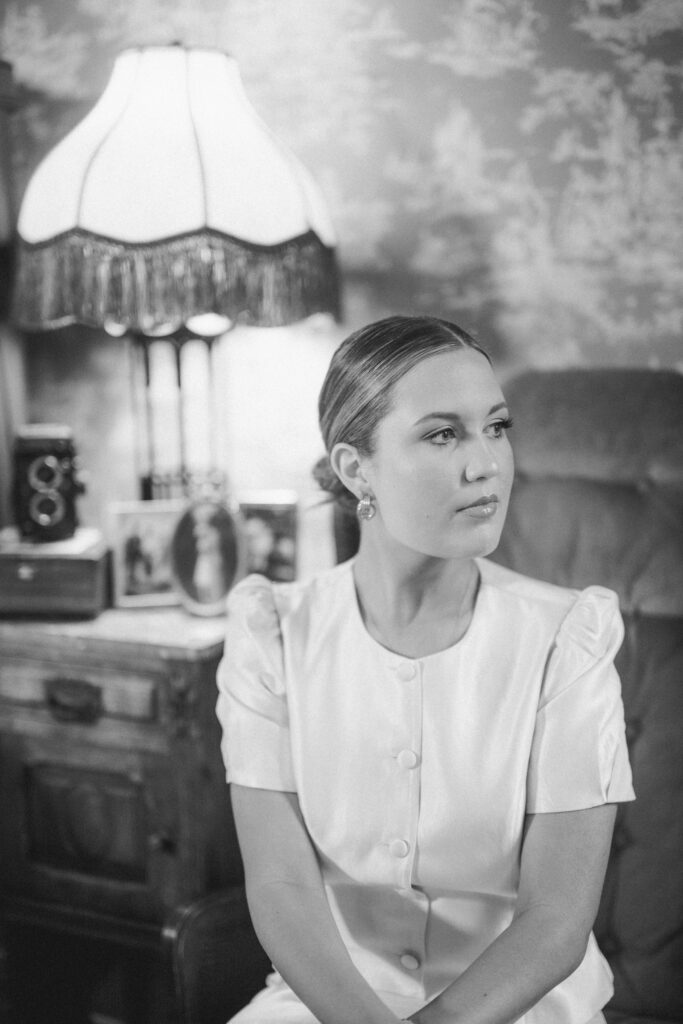 Black-and-white portrait of Lexie Vaccaro sitting near a vintage lamp with framed photographs and a twin-lens reflex camera behind her