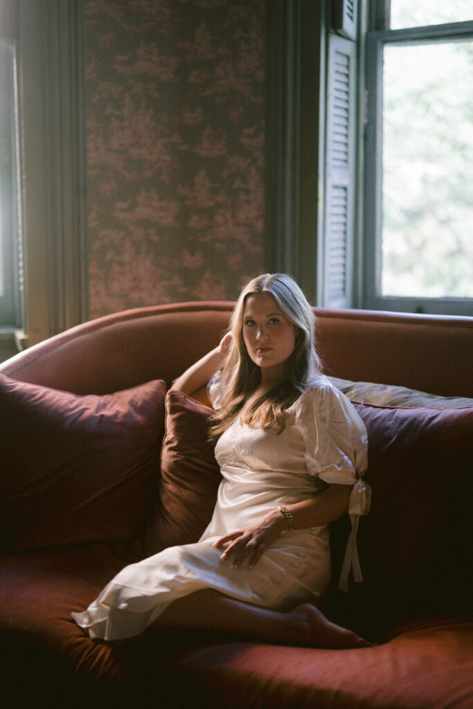 Portrait of Lexie Vaccaro lounging on a velvet sofa in soft window light, wearing a vintage-inspired silk gown