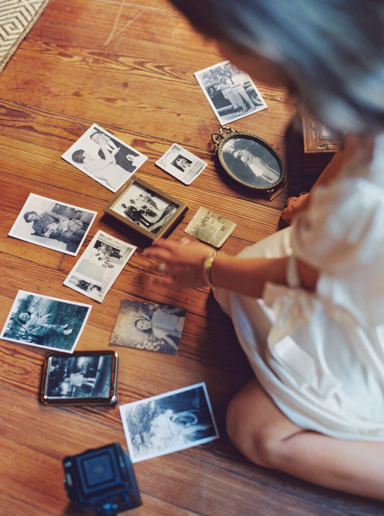 Woman in silk dress sorting through vintage family photographs on wooden floor with a film camera nearby
