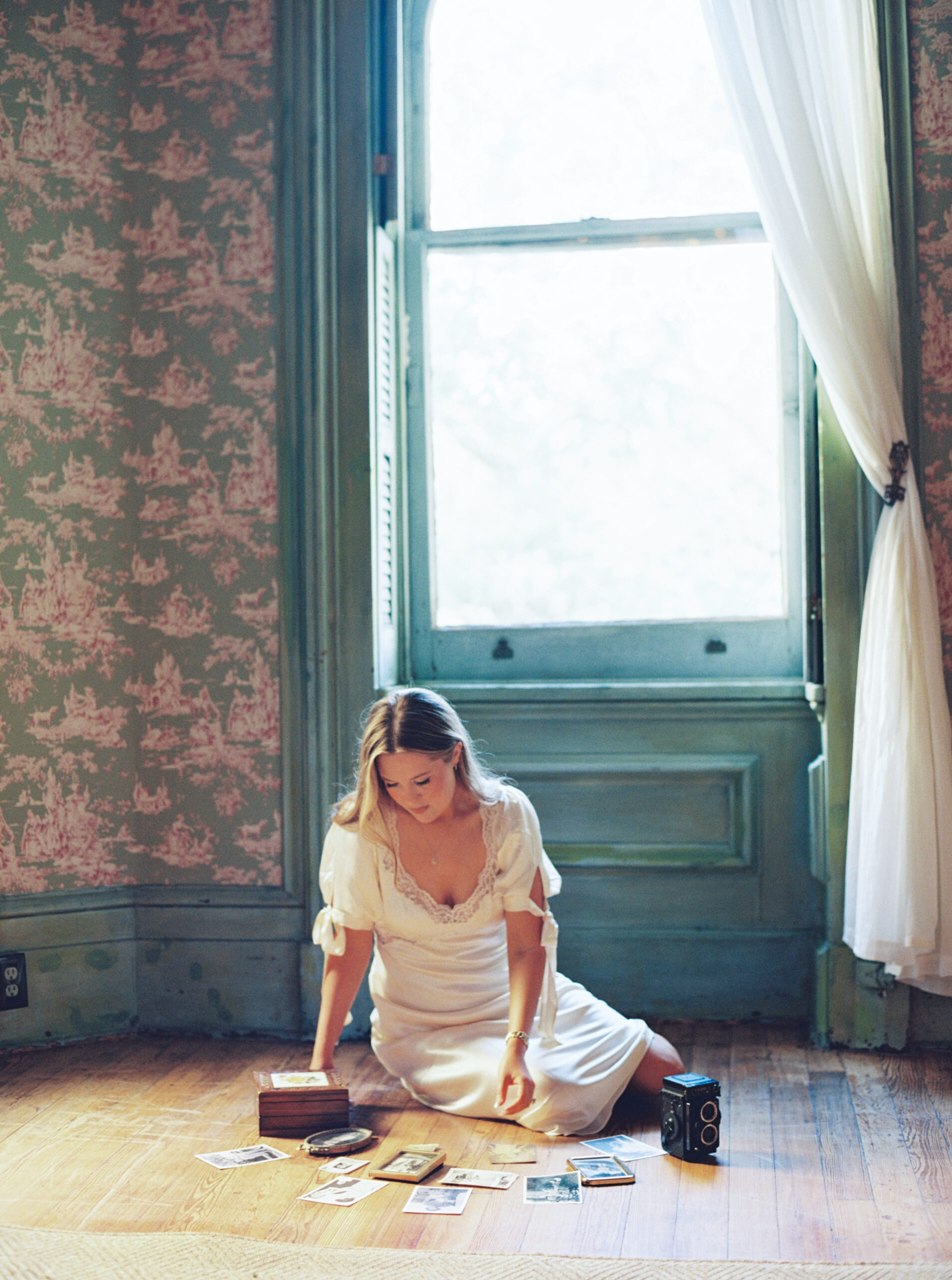 Lexie Vaccaro sitting on a wooden floor in vintage silk Reformation dress, surrounded by old family photographs and a vintage camera