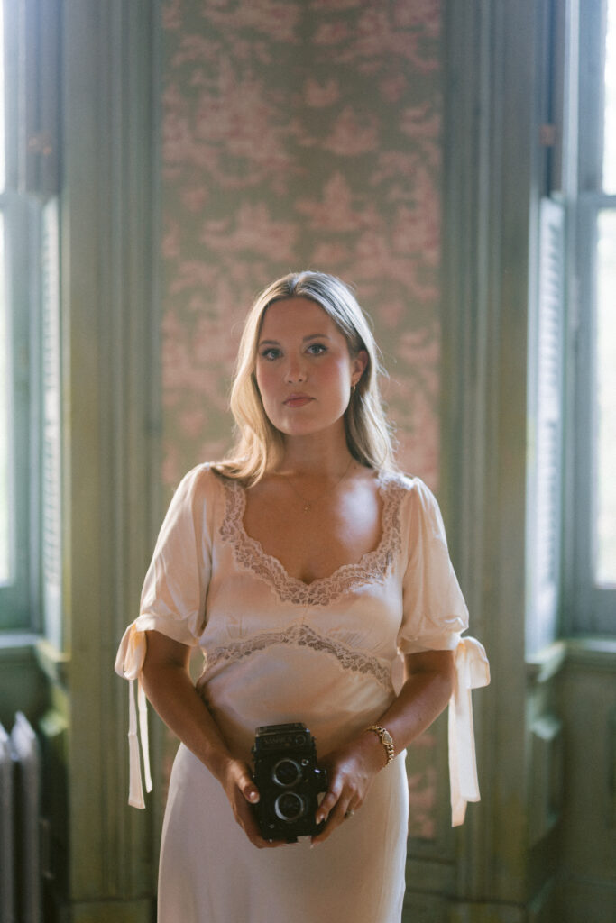 Lexie Vaccaro holding a vintage film camera in soft natural light wearing a silk lace gown