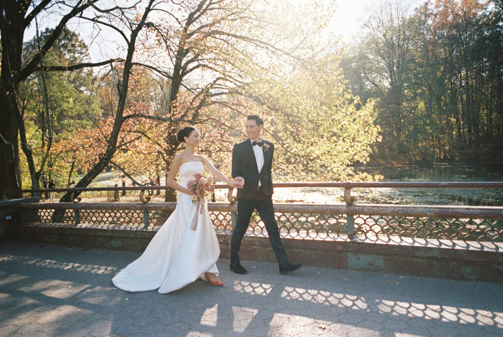 Bride and groom walk hand-in-hand in Central Park, glowing in golden afternoon light with fall foliage in the background