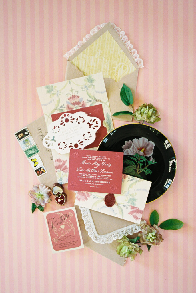 Vintage-inspired wedding invitation suite with red and blush tones, lace details, stamps, and florals on a pink striped background