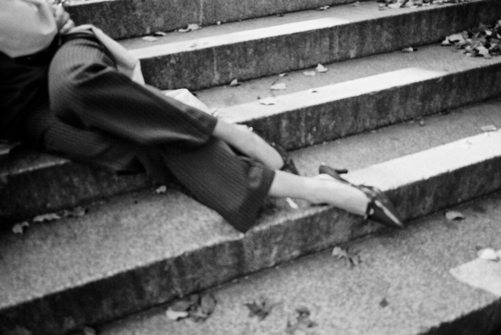Black and white film detail photograph of a woman’s legs in pinstripe trousers and pointed heels, resting on the stone steps at Bethesda Terrace in Central Park, captured by Lexie Vaccaro Photography.