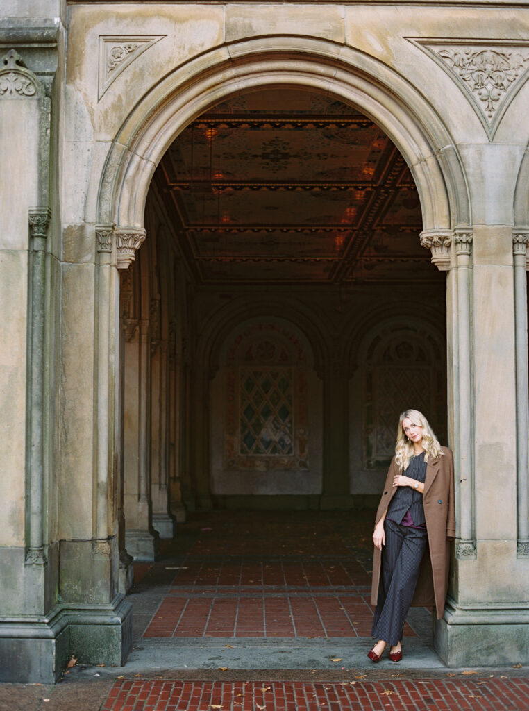 Film photograph of a stylish blonde woman leaning against the stone archway at Bethesda Terrace in Central Park, wearing a brown coat over a pinstripe suit, captured by Lexie Vaccaro Photography.