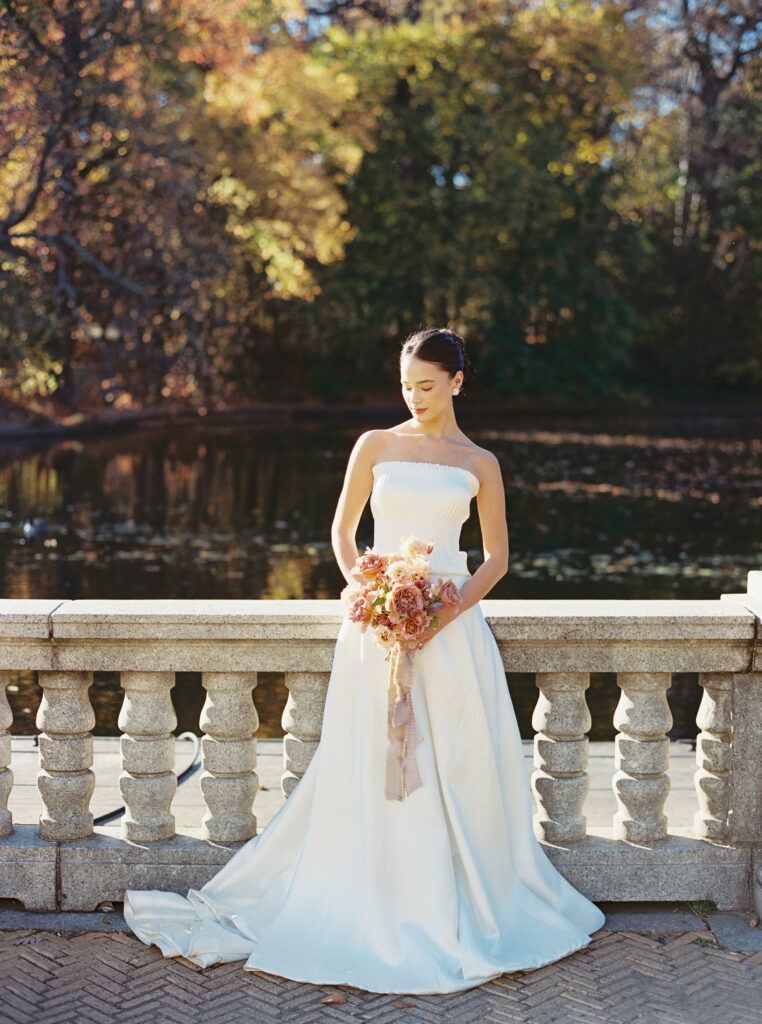 Bride in a strapless white gown holding a blush bouquet, standing beside a lake in Central Park with autumn trees in the background
