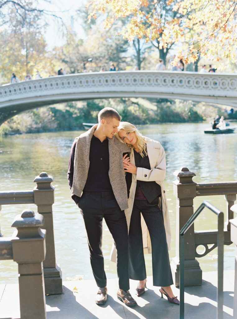 Fall engagement session in Central Park photographed on film by Lexie Vaccaro Photography, featuring a stylish couple standing by Bow Bridge with autumn leaves in the background.