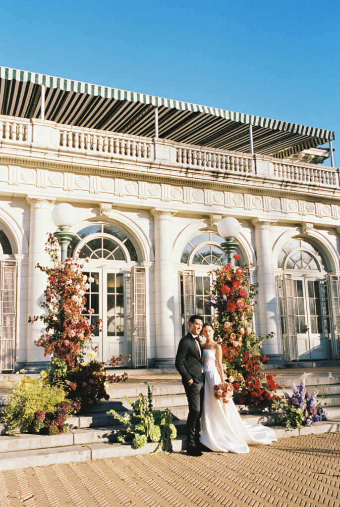 Bride and groom standing on the steps of Purslane at the Boathouse with vibrant floral pillars and architectural backdrop, photographed by Lexie Vaccaro Photography