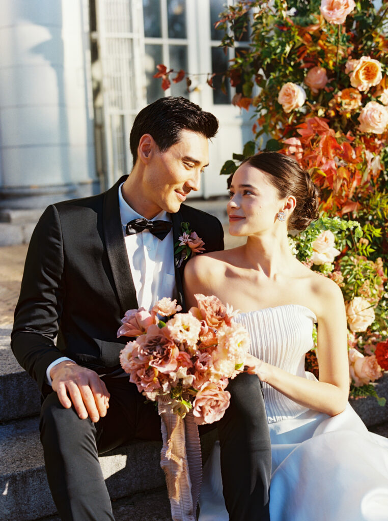 Bride and groom sitting on the steps in front of a floral installation, sharing a soft moment at the Boathouse in Prospect Park, Brooklyn
