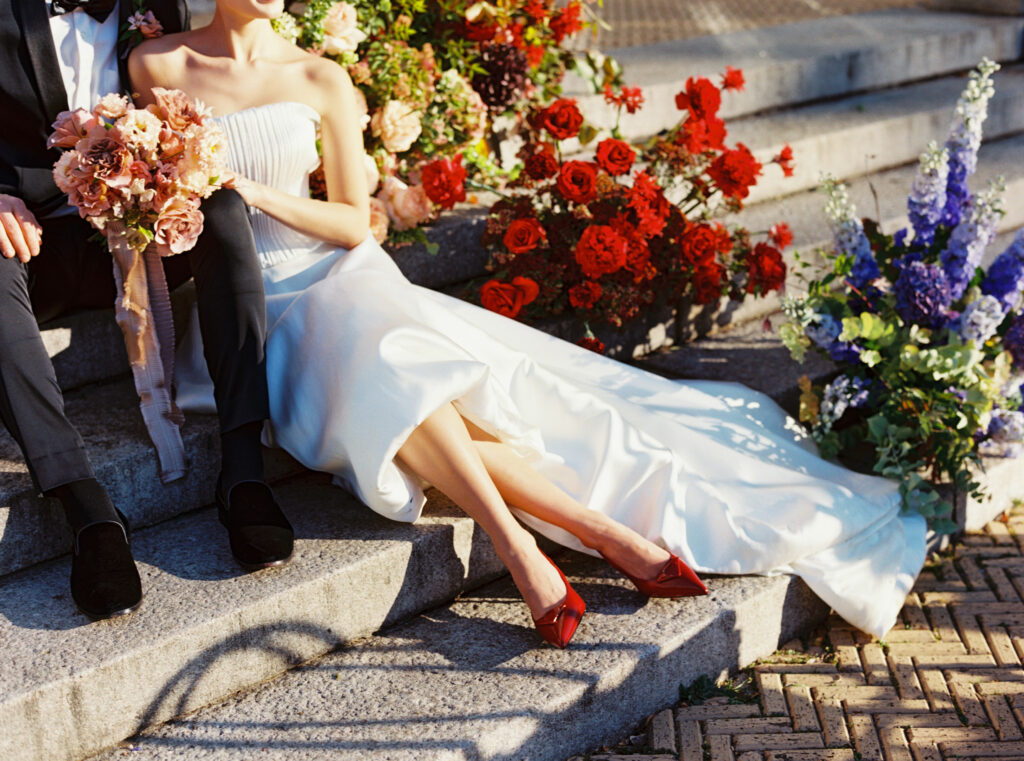 Bride sitting on the stone steps at Purslane at the Boathouse wearing red Prada heels, surrounded by red and purple florals, photographed on film