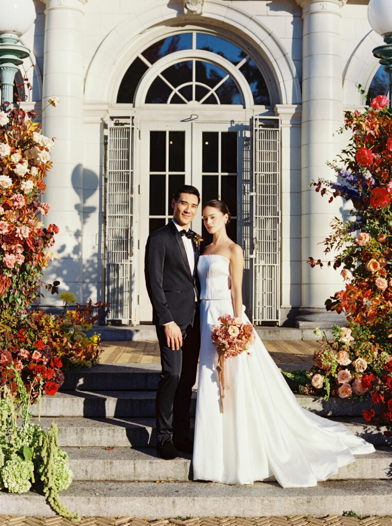 Bride and groom standing on the front steps of the Prospect Park Boathouse surrounded by vibrant floral installations