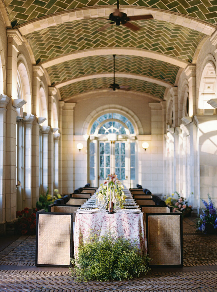 Long wedding reception table with floral centerpieces and tapered candles inside arched conservatory with green tiled ceiling

