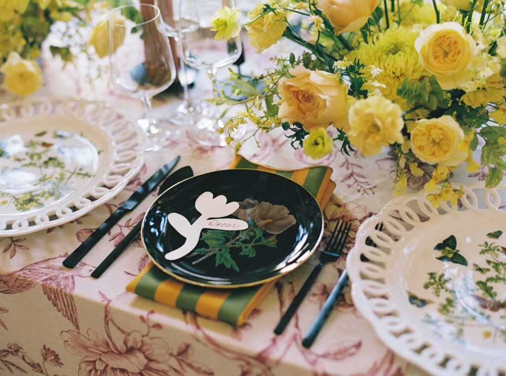 A colorful wedding table setting featuring botanical plates, black and gold flatware, striped napkins, and yellow floral arrangements