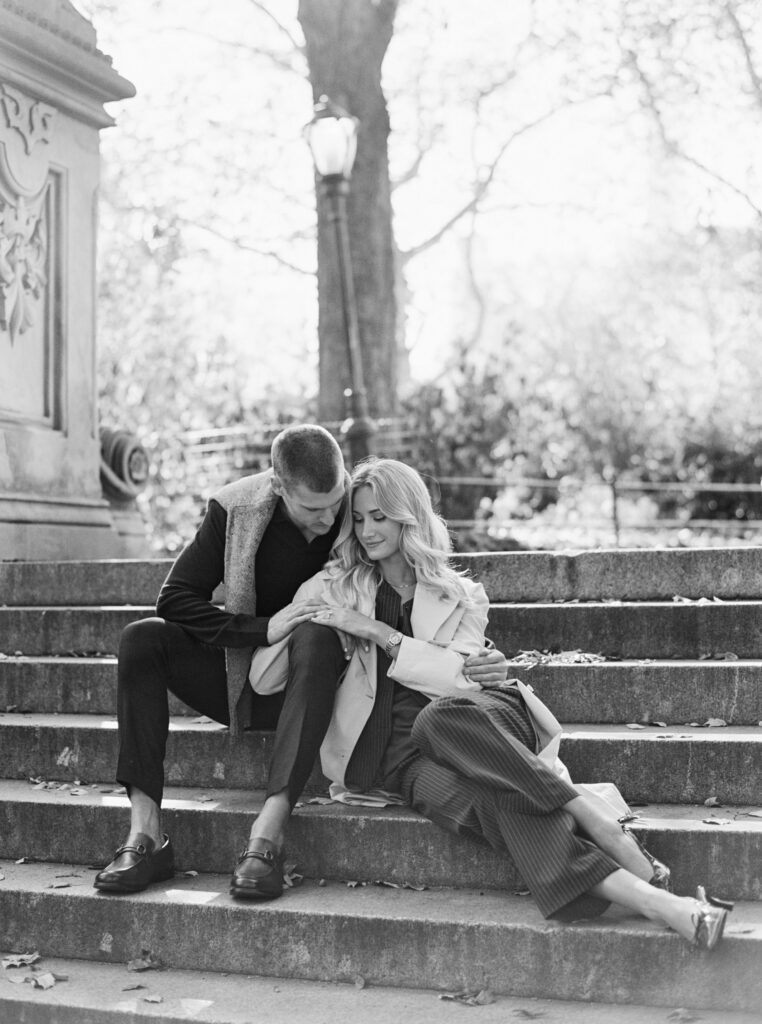 Black and white film photograph of a stylish couple sitting on the Bethesda Terrace steps in Central Park during fall, wearing tailored coats and loafers.