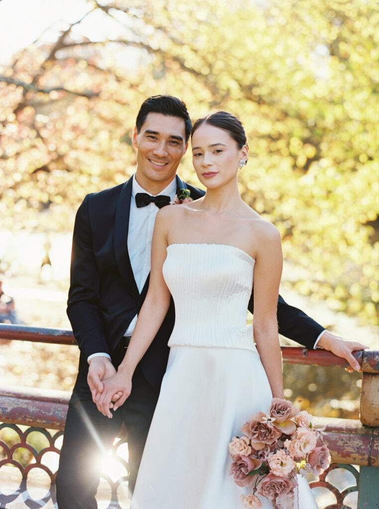 Bride and groom holding hands on a bridge in Prospect Park with golden fall light and a soft mauve bouquet, photographed by Lexie Vaccaro Photography
