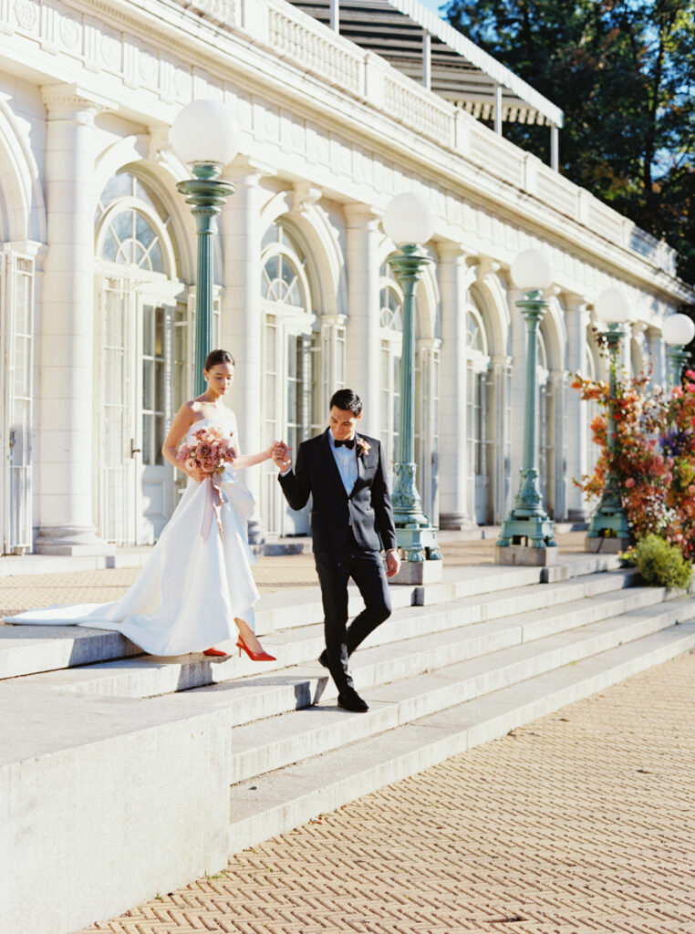 Bride and groom walk down the steps of the Beaux-Arts Boathouse in Prospect Park, Brooklyn, with elegant architecture and floral installation in the background