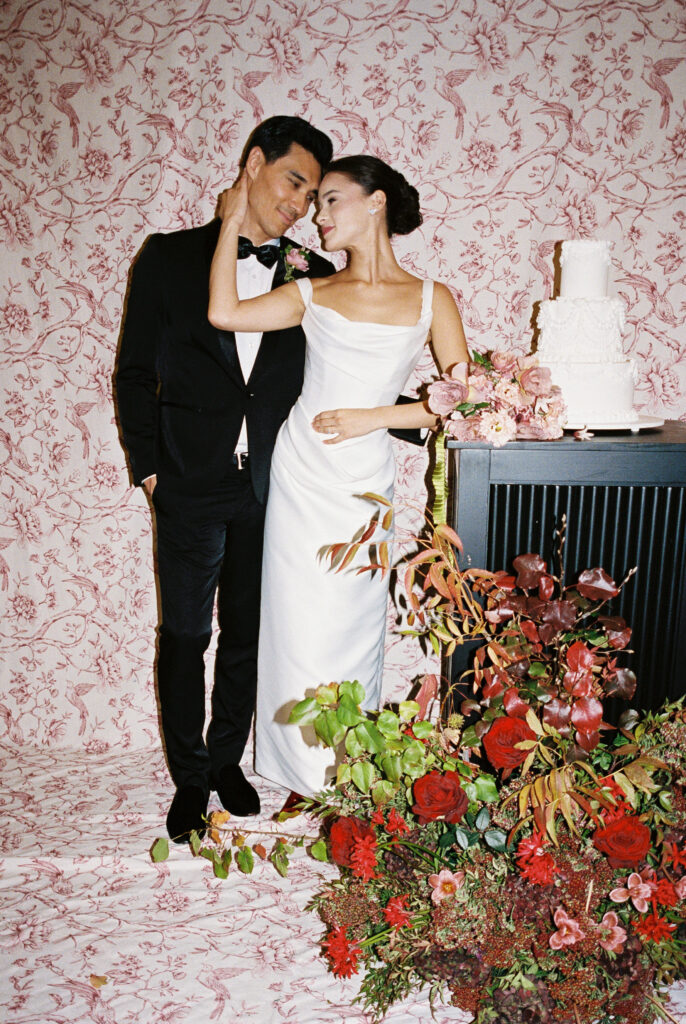 Bride and groom sharing an intimate moment in front of a vintage floral wallpaper backdrop with a white wedding cake and lush floral arrangement