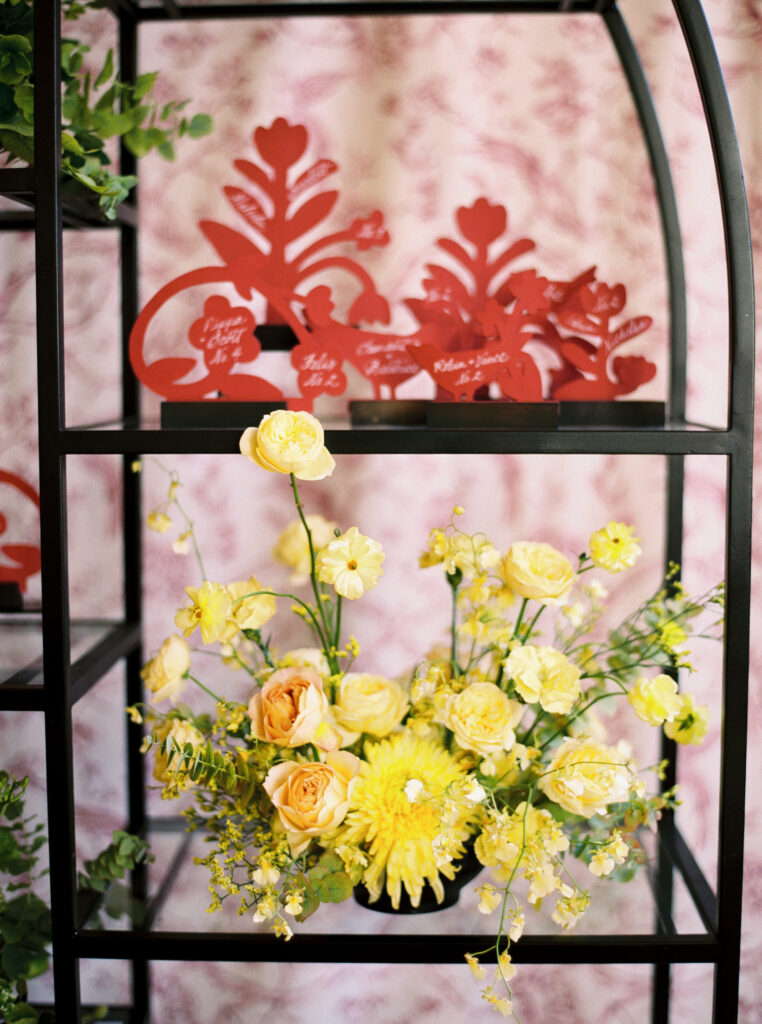 Red folk-art inspired escort cards displayed on black shelving with yellow floral arrangement below, photographed on film